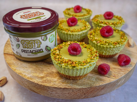 Po lewej stronie znajduje się krem pistacjowy Pistachio bez dodatku cukru 200g, a obok niego deserki: Mini serniczki pistacjowe ozdobione malinami.